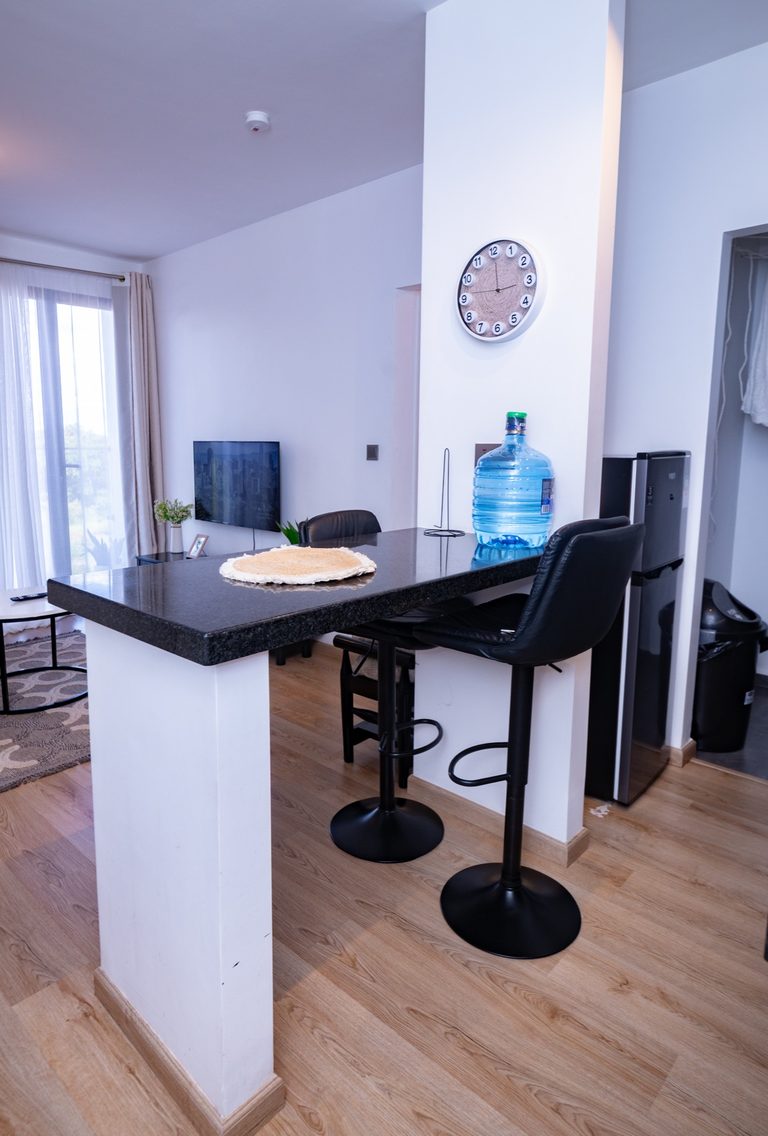 Kitchen breakfast bar with two black stools, a wall clock and a water dispenser, TV visible nearby, The Graphite, Tatu City