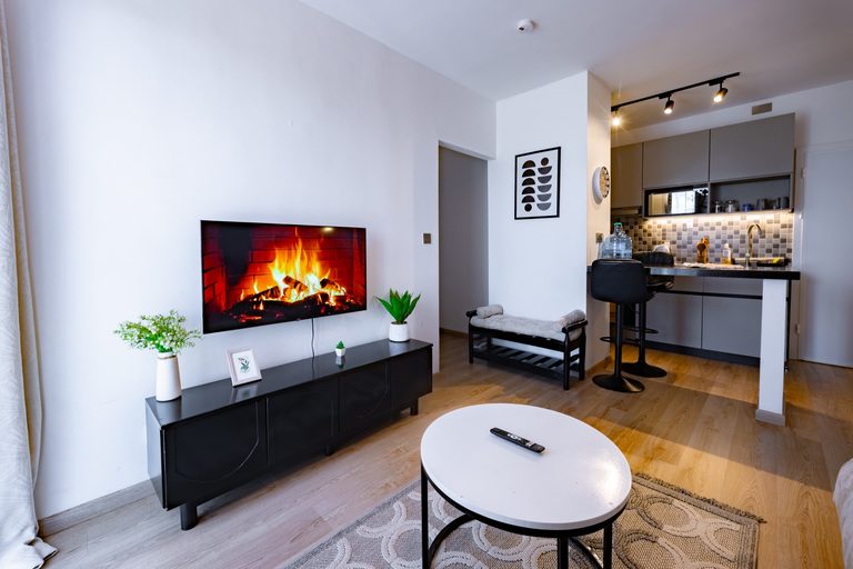 TV wall playing a fireplace video, black media console and a bench with bolster cushions, kitchen beyond, The Graphite, Tatu City